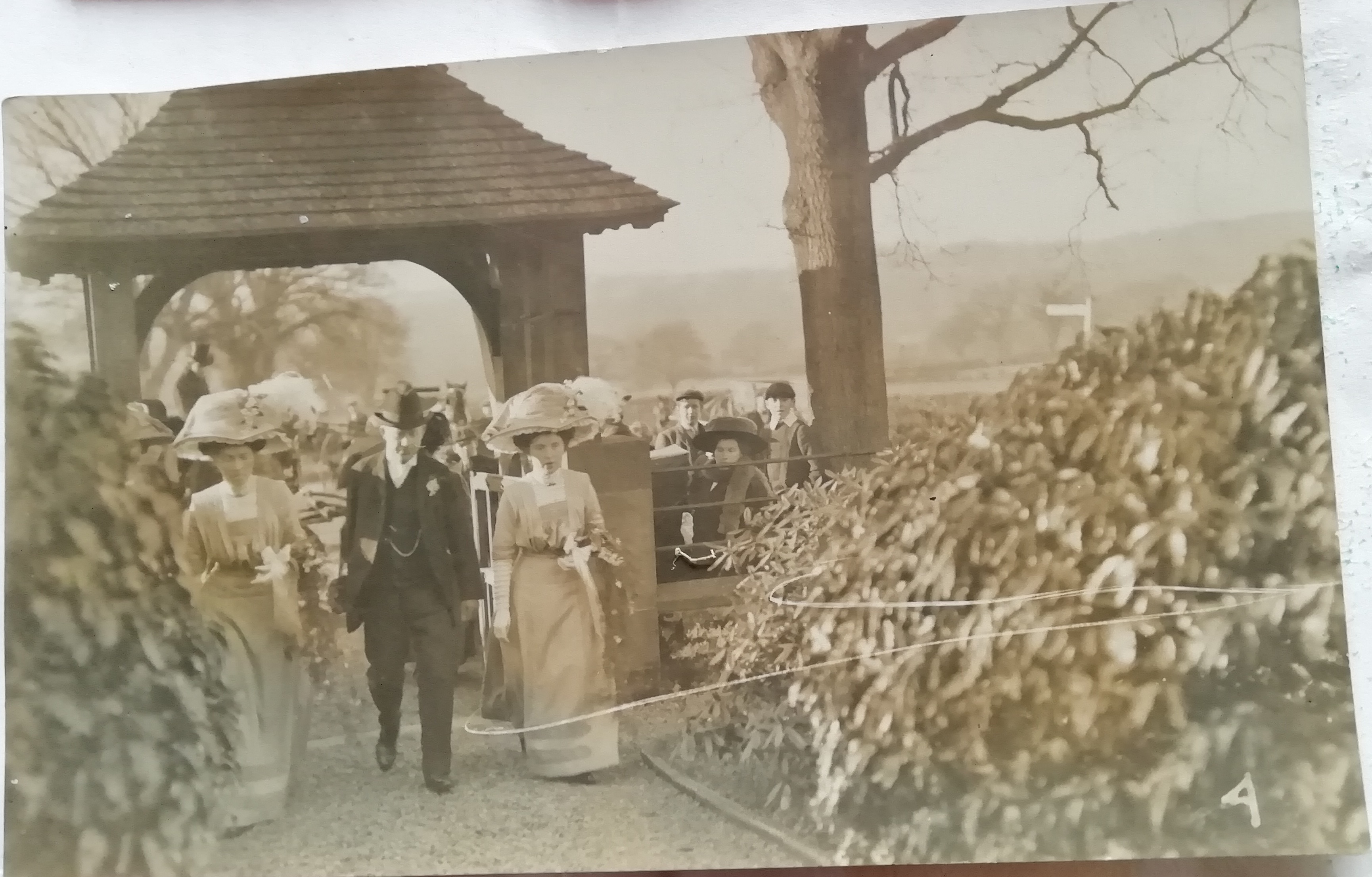 Black and white photograph of wedding party by lychgate. 2 brides with gentleman in-between. 1911.